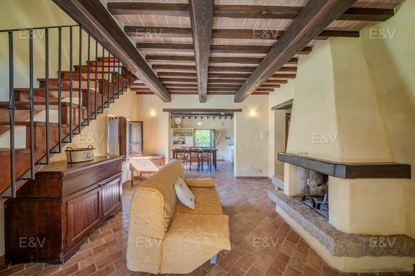 Interior view of a living room with a fireplace, sofa, and staircase. The room has exposed wooden beams and terracotta tile flooring.