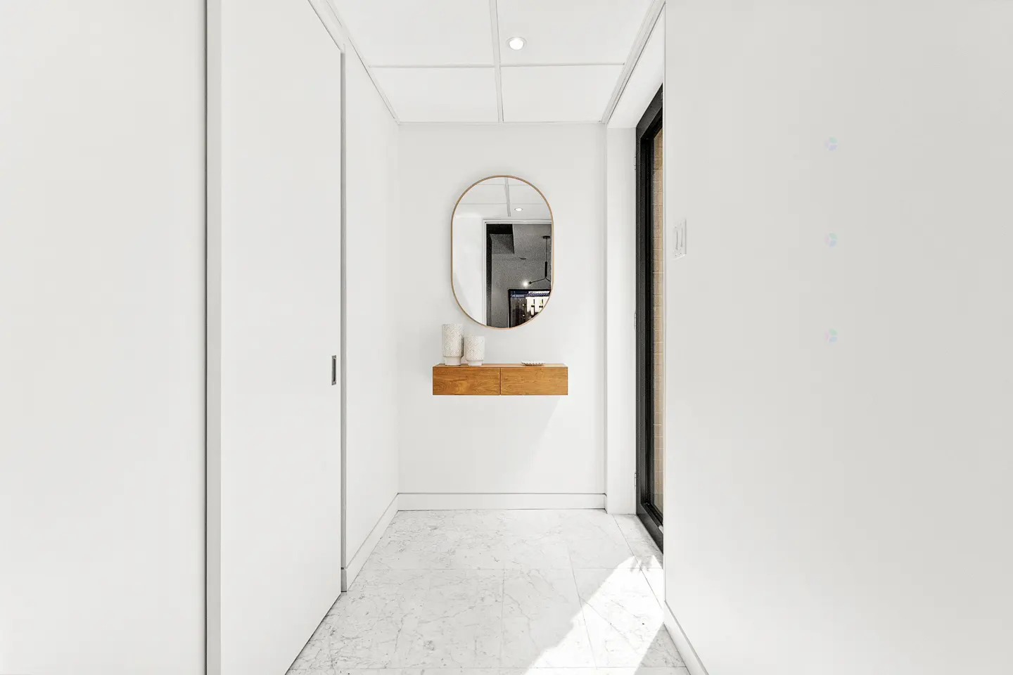 Bright hallway with white walls, marble floor, and a wood floating shelf with an oval mirror above it.