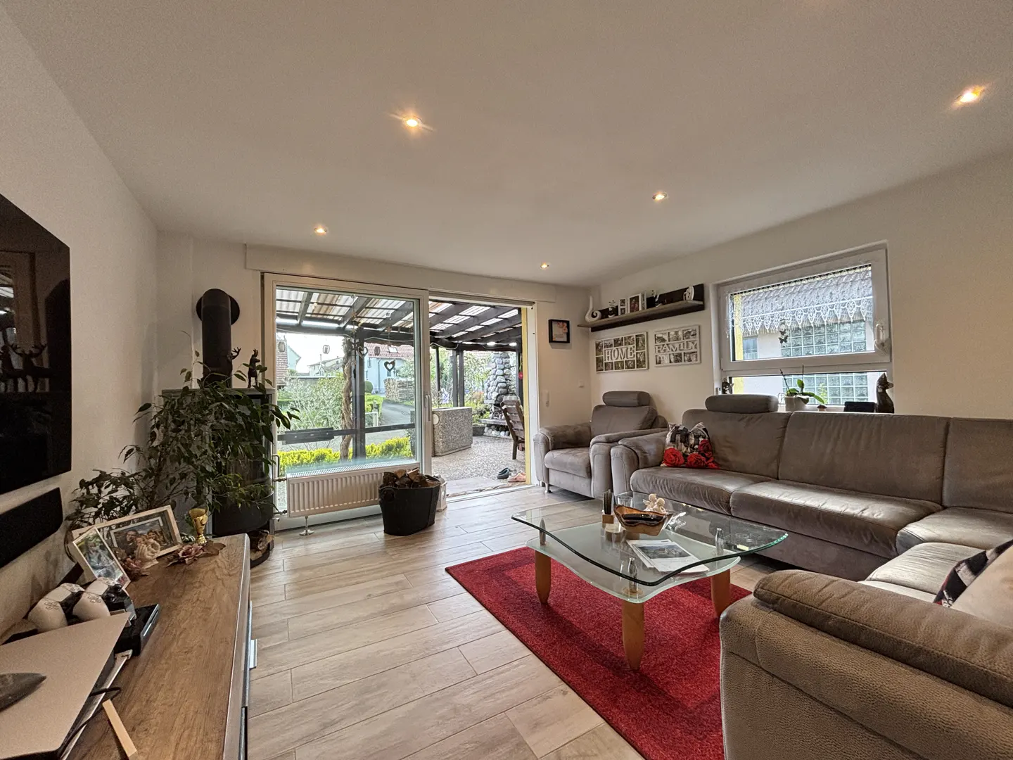 Bright living room with a grey sectional sofa, glass coffee table on a red rug, wood-look tile floors, and patio access.