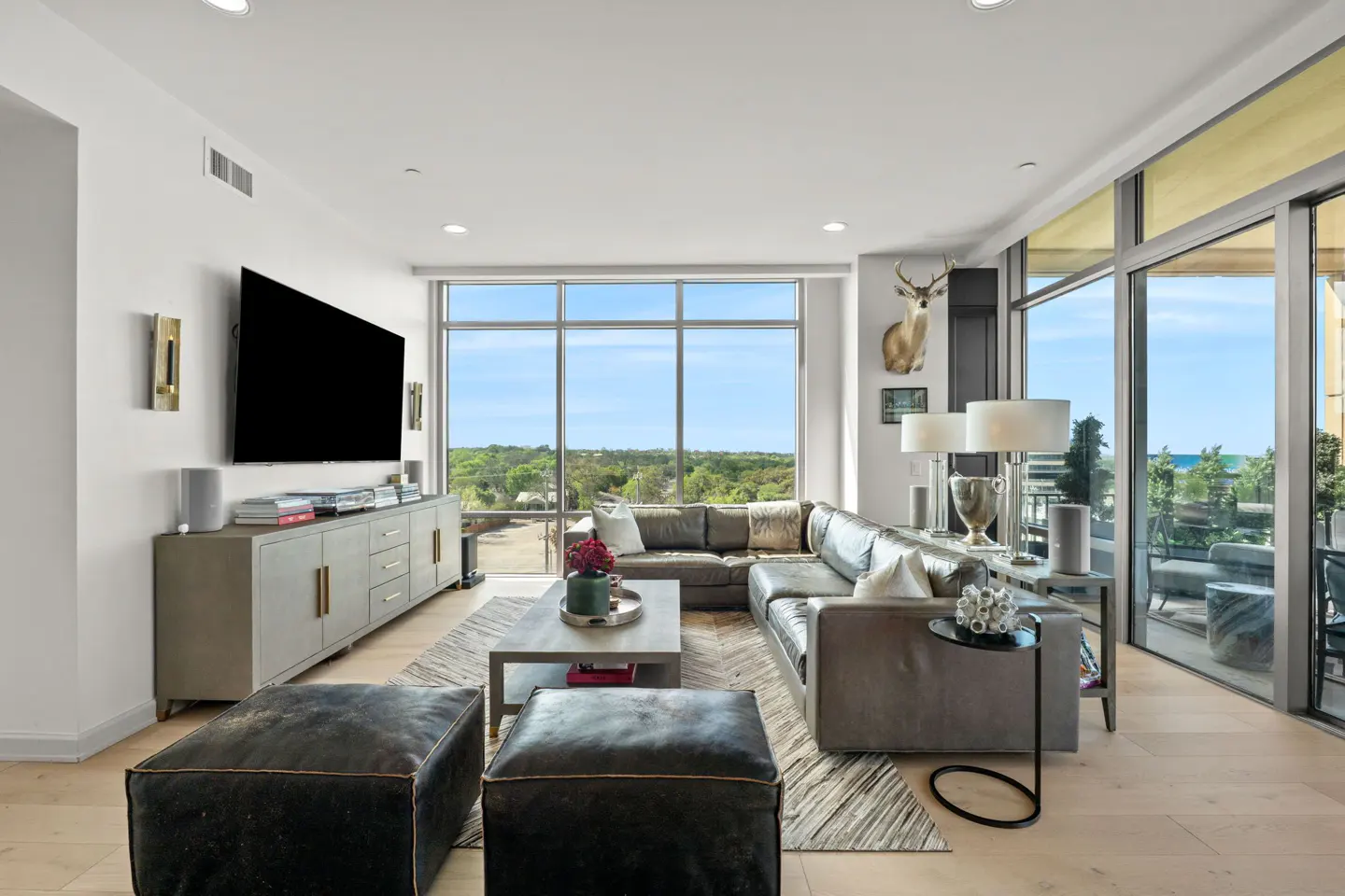 Bright living room with a gray sectional sofa, two black ottomans, and a large window with a view of trees and blue sky.