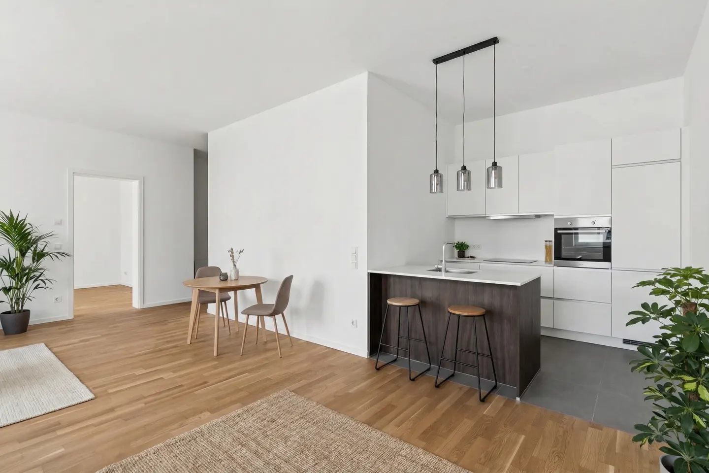 Bright, modern apartment interior with wood floors, white walls, and a kitchen island with stools. A round table and chairs sit near a doorway.
