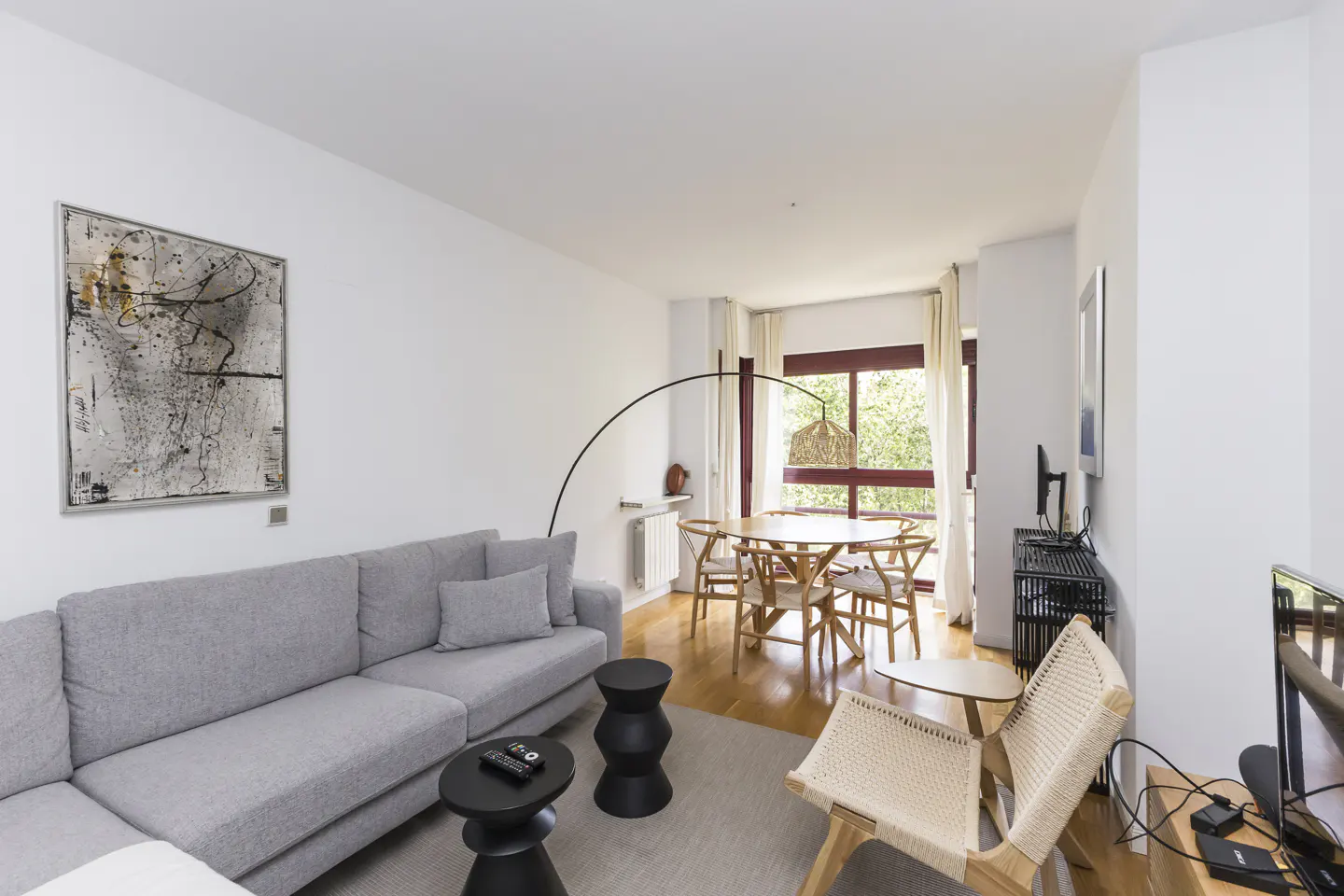 Bright living room with a gray sectional sofa, black side tables, and a round wooden dining table near a window. An abstract painting hangs on the white wall.