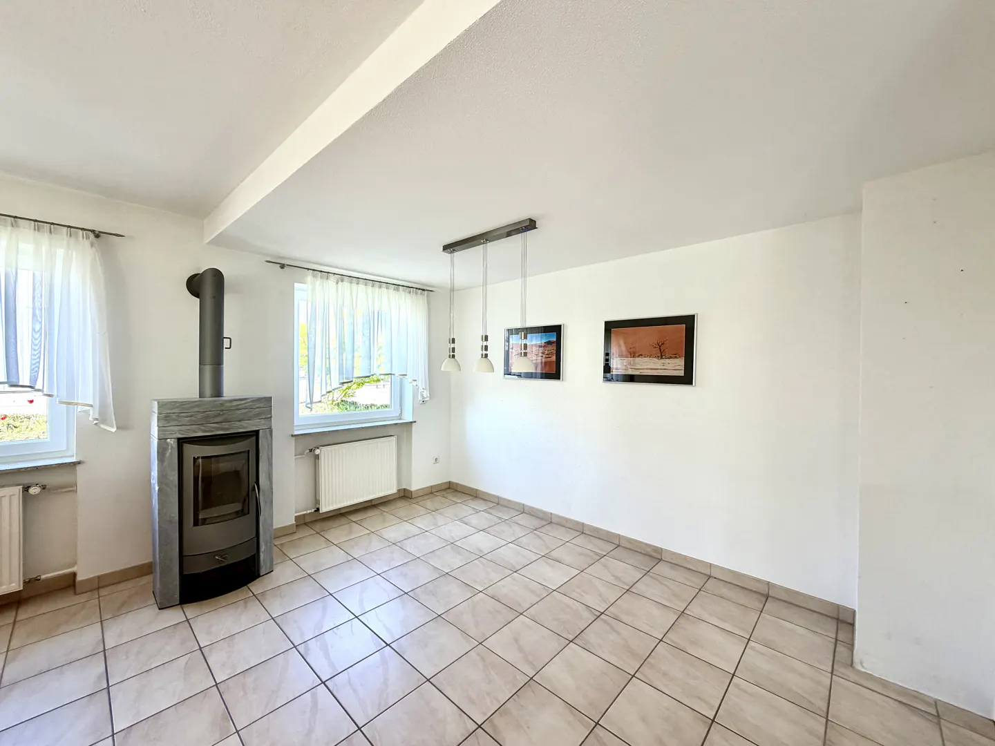 Bright room with tile floor, white walls, and a wood-burning stove. Two windows with sheer curtains, two framed pictures, and a pendant light fixture.