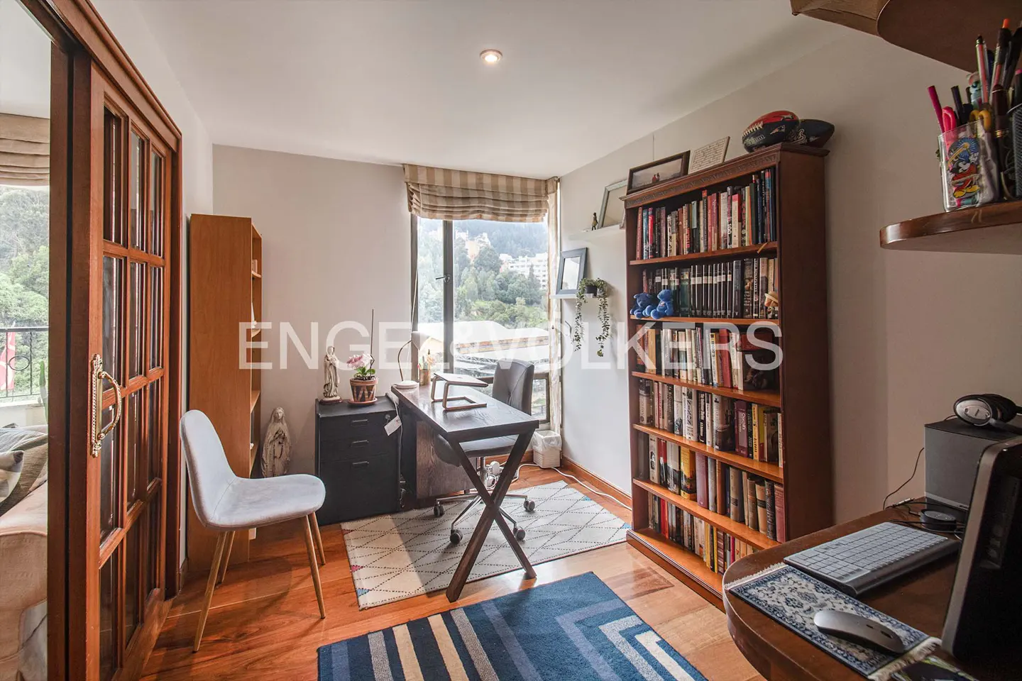 A bright home office with a wooden floor, a desk by the window, and a tall bookcase filled with books.