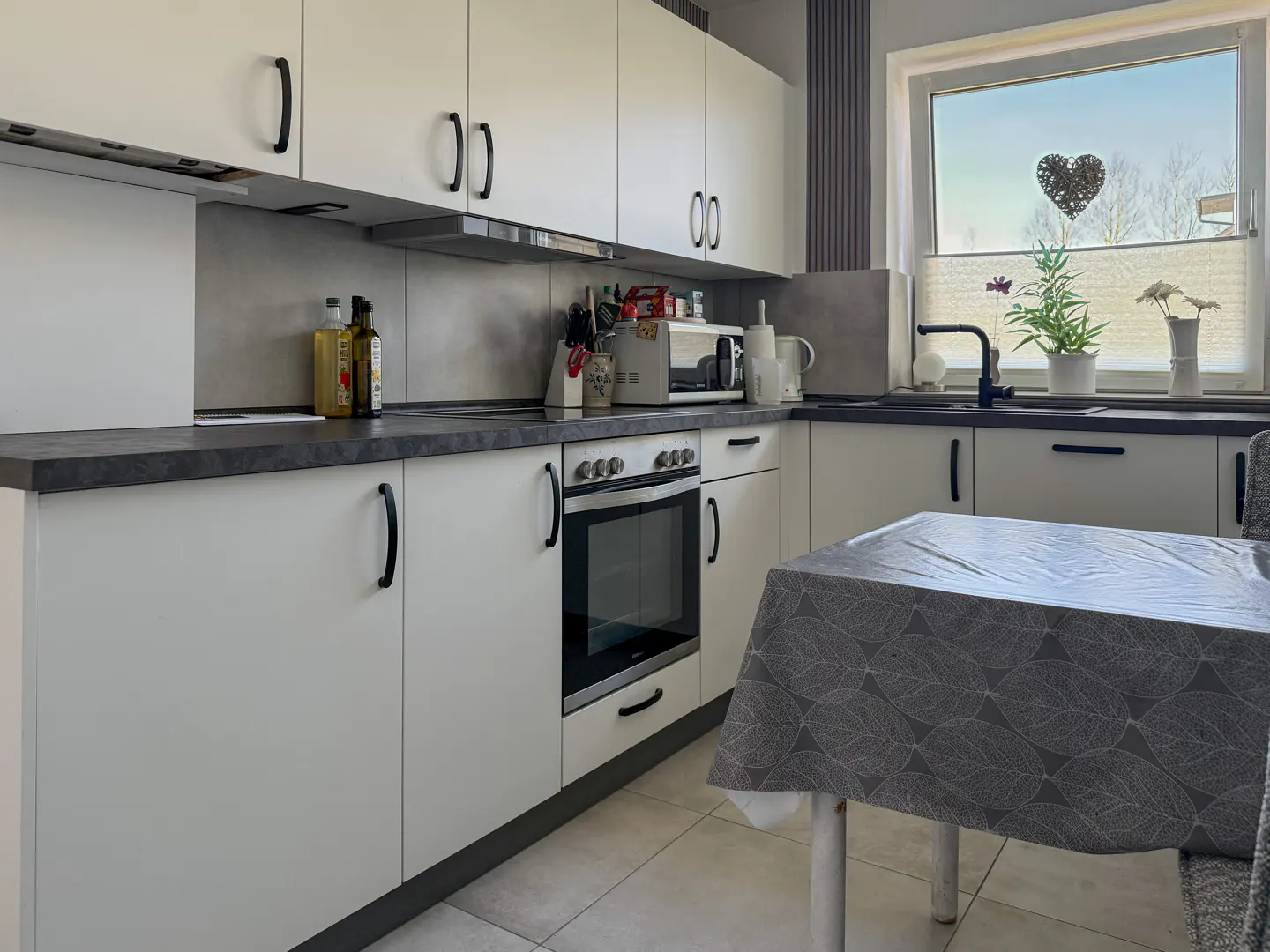 A bright kitchen with white cabinets, black handles, and a dark countertop. A table with a gray leaf-patterned tablecloth is in the foreground. A window with a heart decoration is visible.