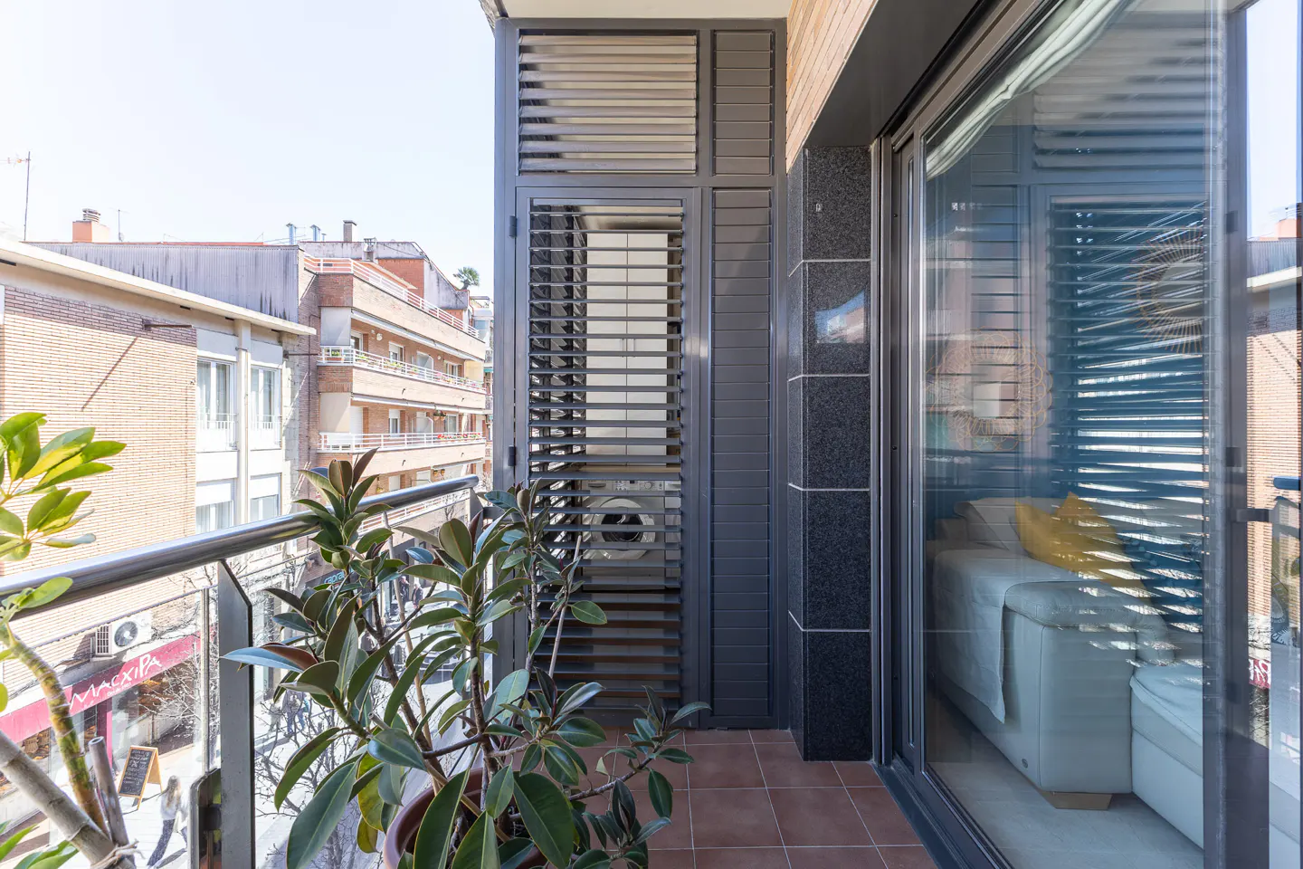 Balcony view with plants, gray shutters, and sliding glass doors. A washing machine is visible through the shutters. City buildings in the background.