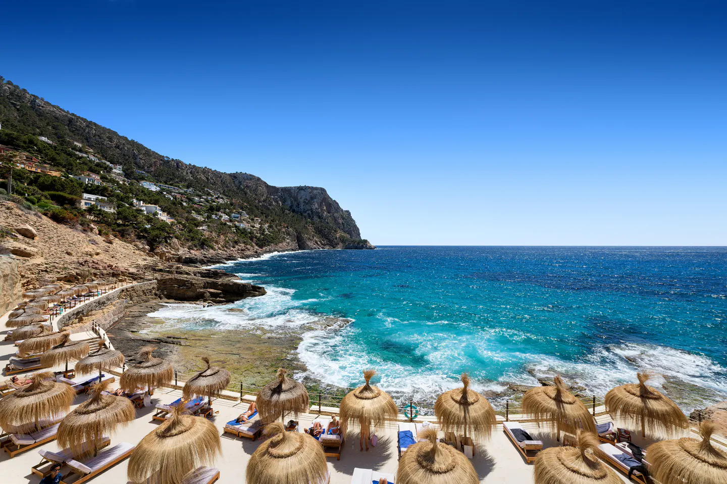 Beachfront resort with straw umbrellas and lounge chairs overlooking turquoise water and a rocky coastline under a clear blue sky.