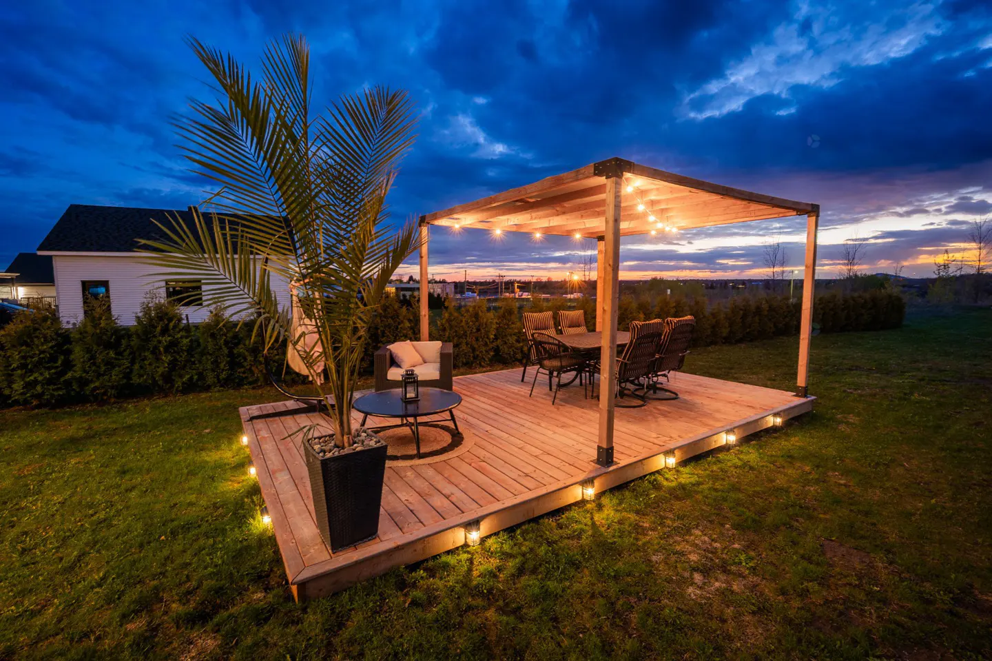 Outdoor patio with wooden pergola, string lights, table, chairs, and potted palm tree at dusk. Lawn and house in background.