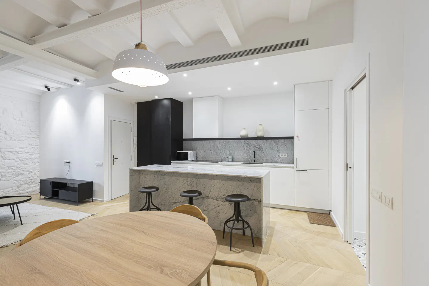 Bright, modern kitchen and living area with white walls, wood floors, and a marble island with stools. A round wood table sits in the foreground.