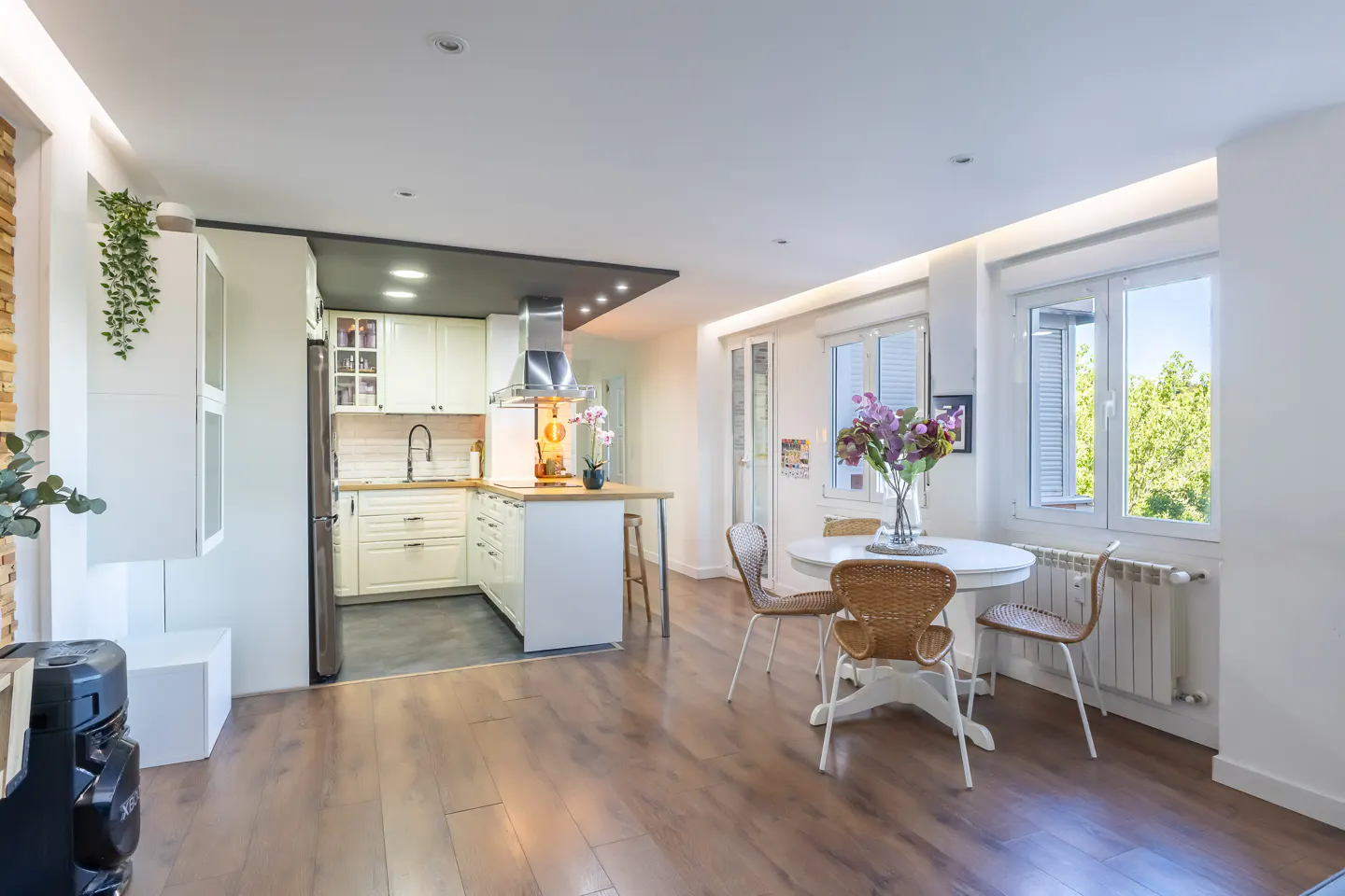 Bright, open-concept apartment with wood floors, white kitchen cabinets, and a round dining table with wicker chairs near a window.