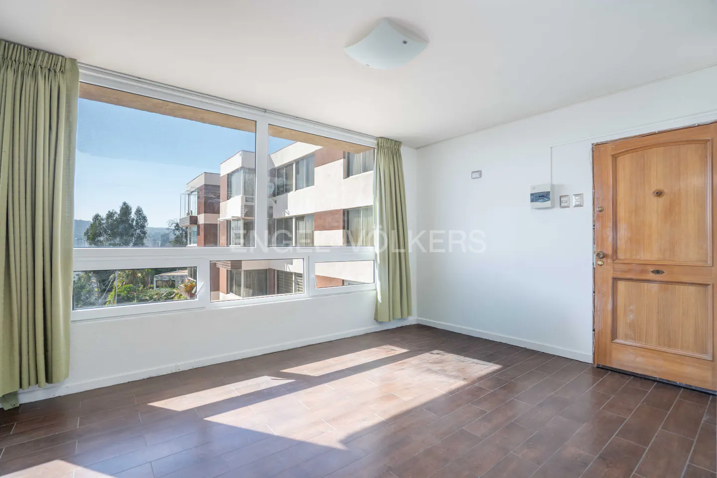 A bright, empty room with hardwood floors, a large window with a view, and a wooden door.