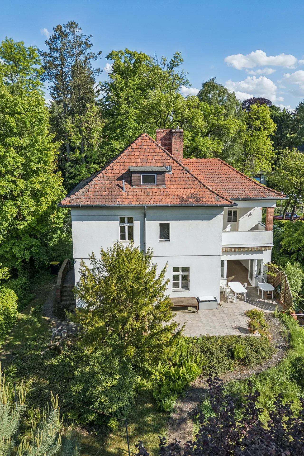 White two-story house with a red tile roof, surrounded by lush green trees, featuring a patio with outdoor furniture.