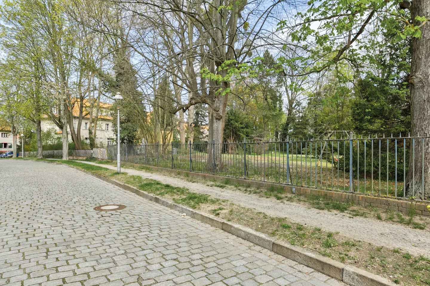 Cobblestone street view with a metal fence bordering a grassy area with trees and a building in the background.