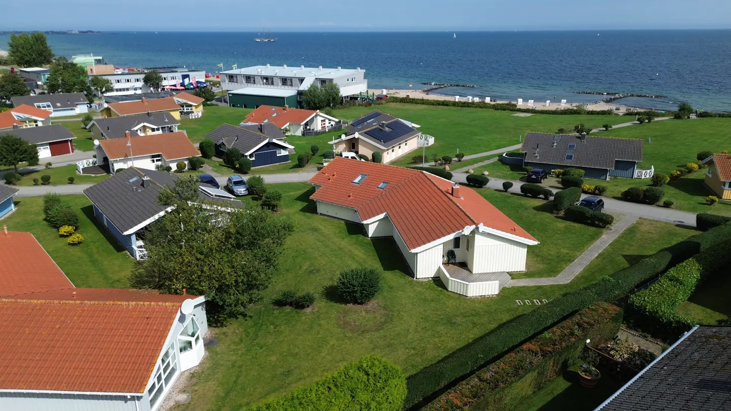 Aerial view of a coastal neighborhood with colorful houses, green lawns, and the blue sea in the background.
