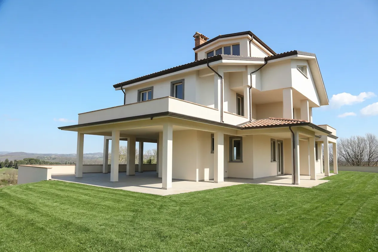 A modern, two-story beige house with a green lawn under a clear blue sky. The house has a covered patio with white pillars.
