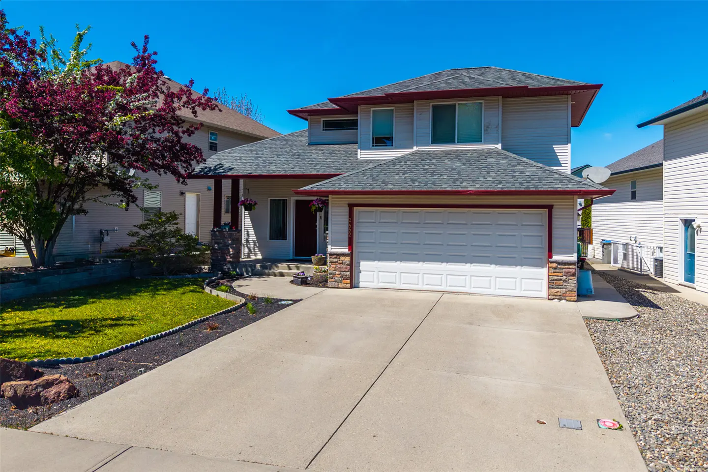 Two-story house with gray siding, gray roof, and white garage door. A tree with purple leaves is on the left. Concrete driveway. Blue sky.
