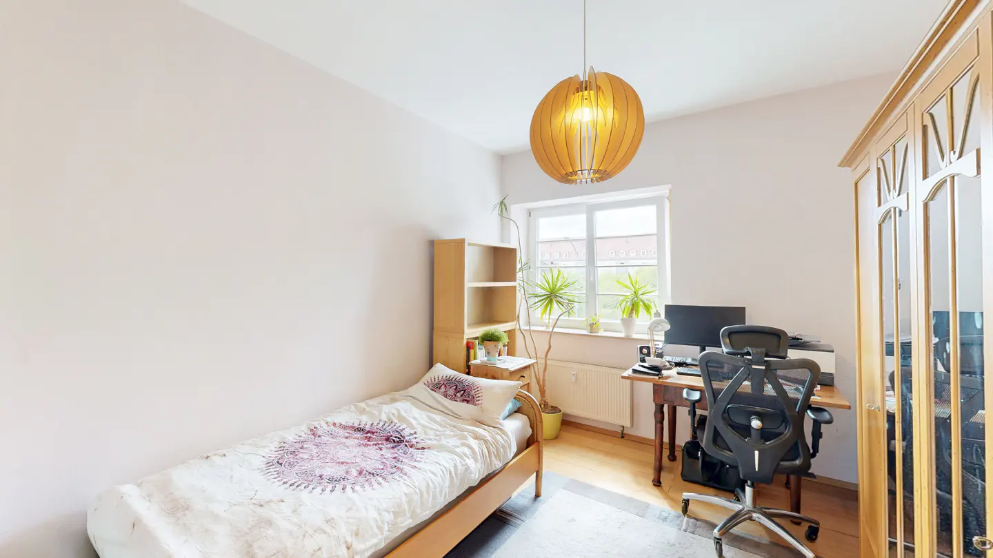 A bright bedroom with a single bed, desk, computer, and wooden wardrobe. A unique wooden pendant light hangs from the ceiling.