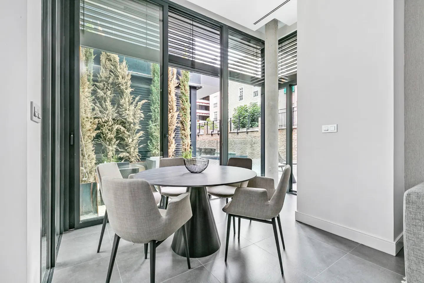 A modern dining area with a round table, gray chairs, and large windows overlooking greenery.