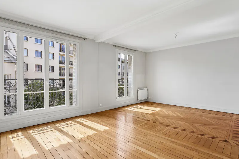 Bright, empty room with parquet floors, white walls, and large windows. Outside, a building and greenery are visible.