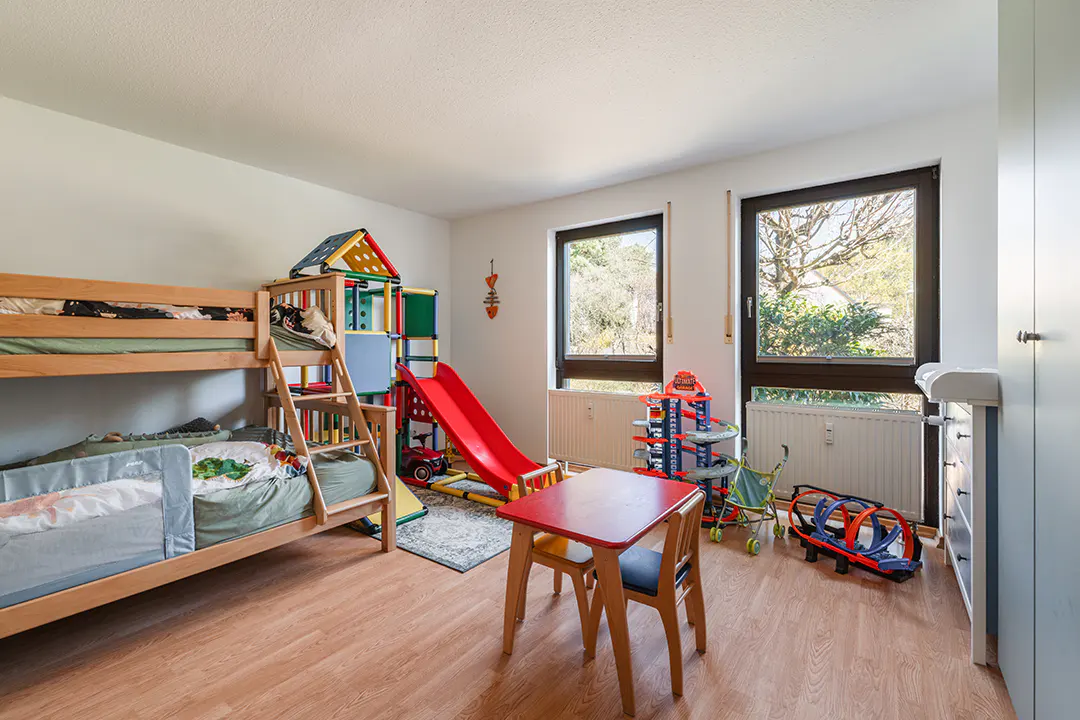 A child's bedroom with a wooden bunk bed, red slide, table, and toys. Two windows provide natural light.