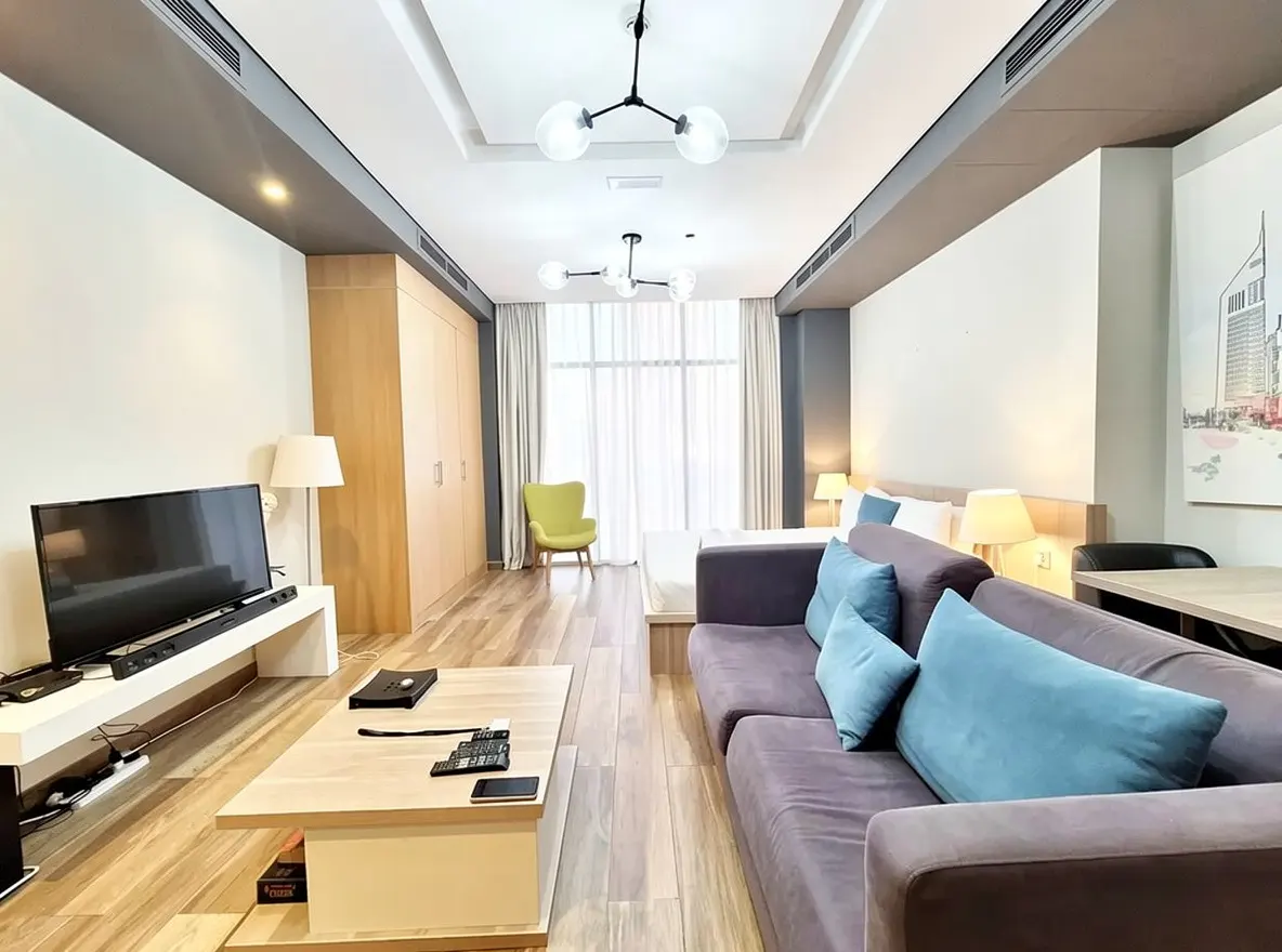 A modern studio apartment with a gray sofa, blue pillows, TV, and a light wood floor. A bed and a green chair are in the background.