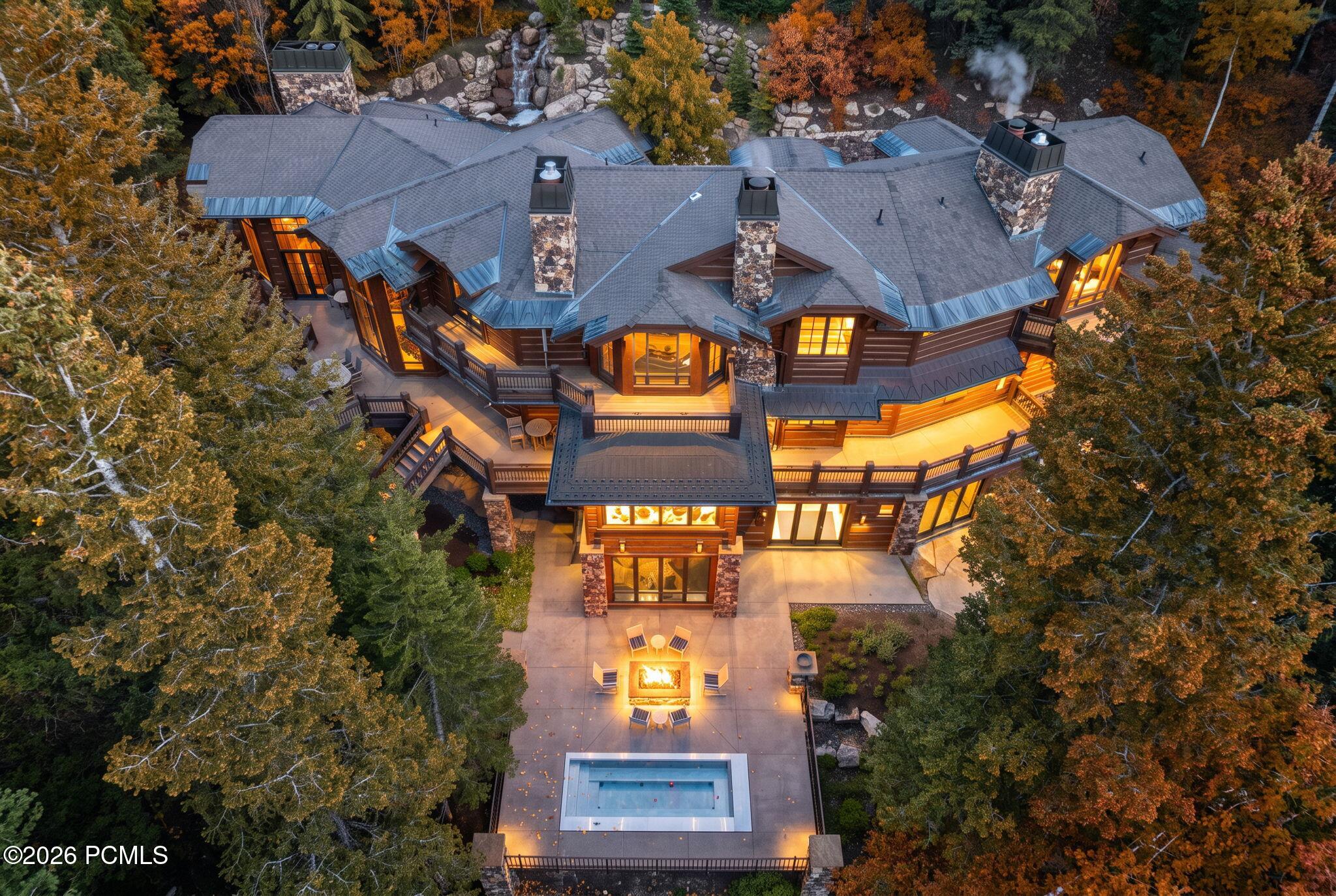 Aerial view of a luxury mountain home with a stone patio, fire pit, and pool, surrounded by autumn trees at dusk. [AI-generated text]
