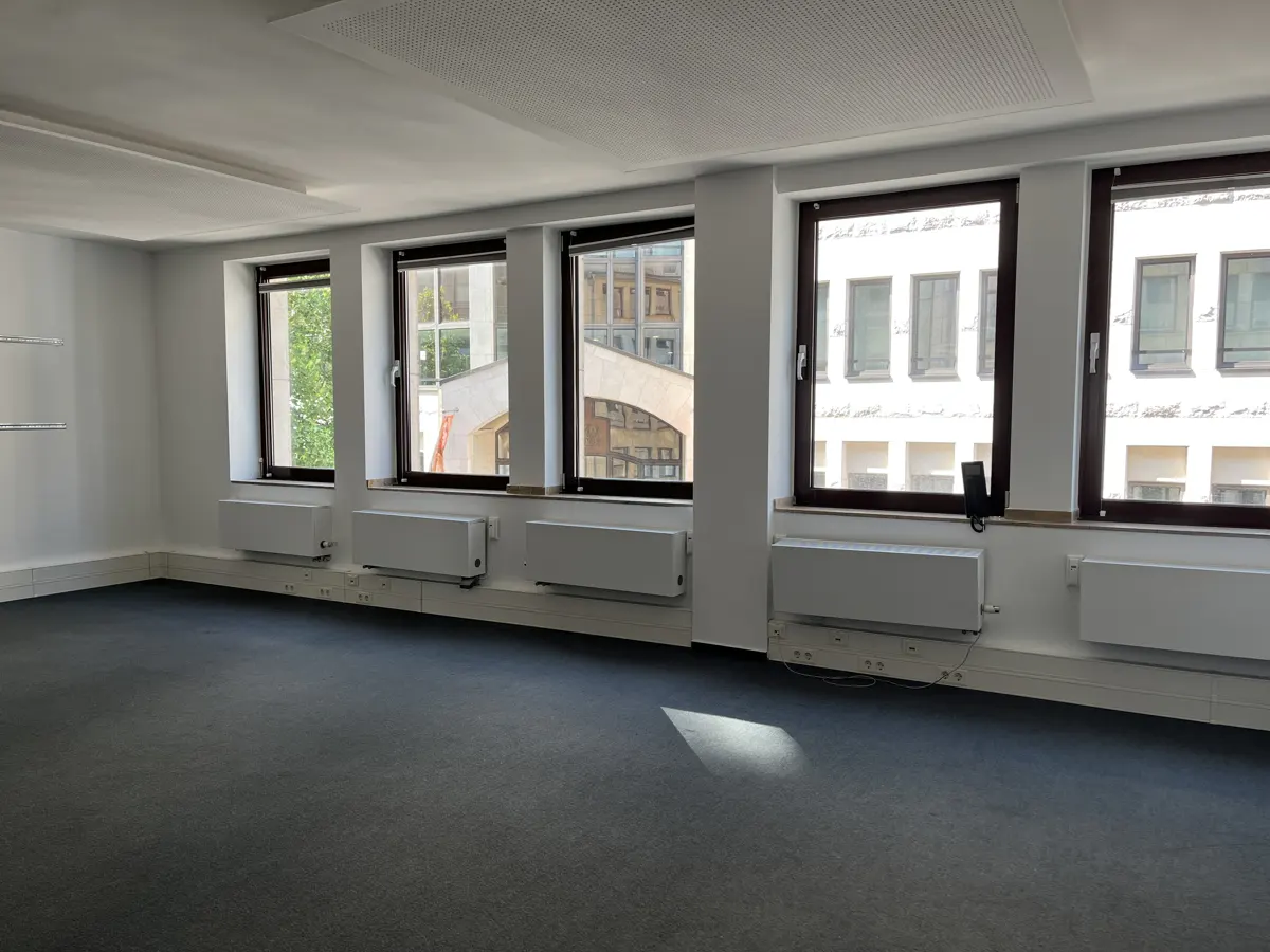 Bright, empty office space with gray carpet, white walls, and five windows showing buildings outside.