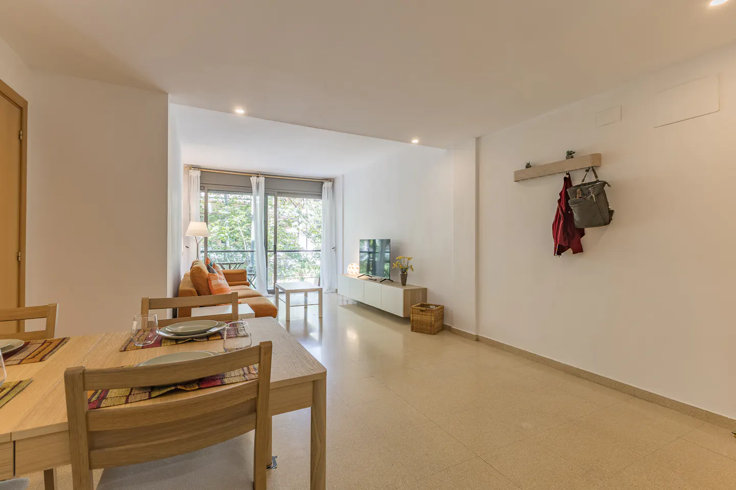 Bright open-plan living room with a dining table, orange sofa, TV console, and a wall-mounted coat rack with a bag.