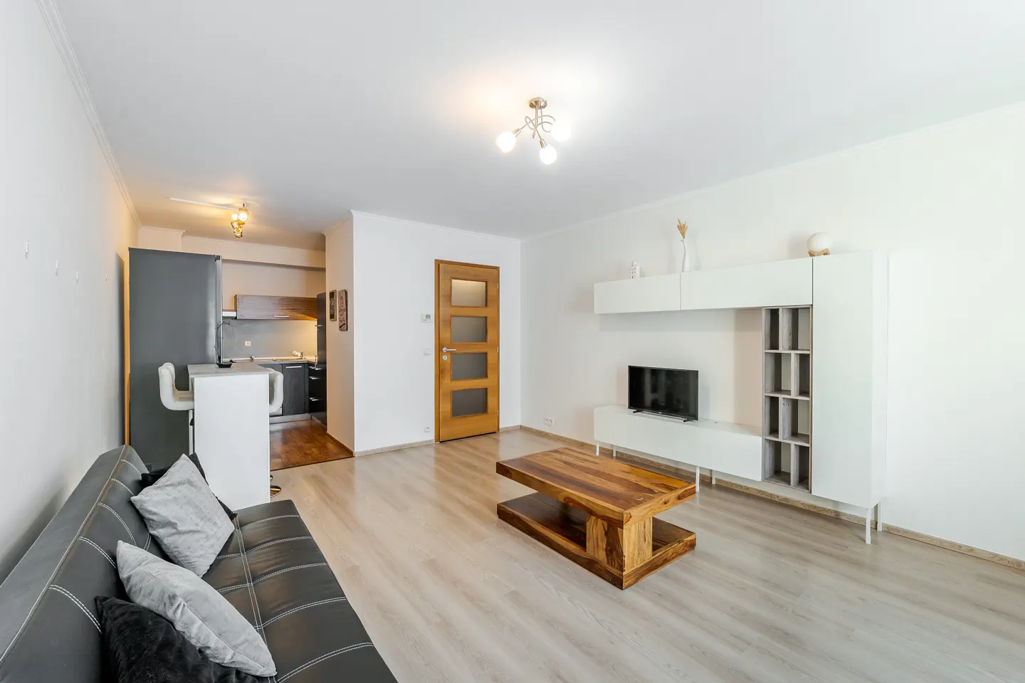 Bright living room with a black sofa, wooden coffee table, and white entertainment center. Kitchen visible in the background.