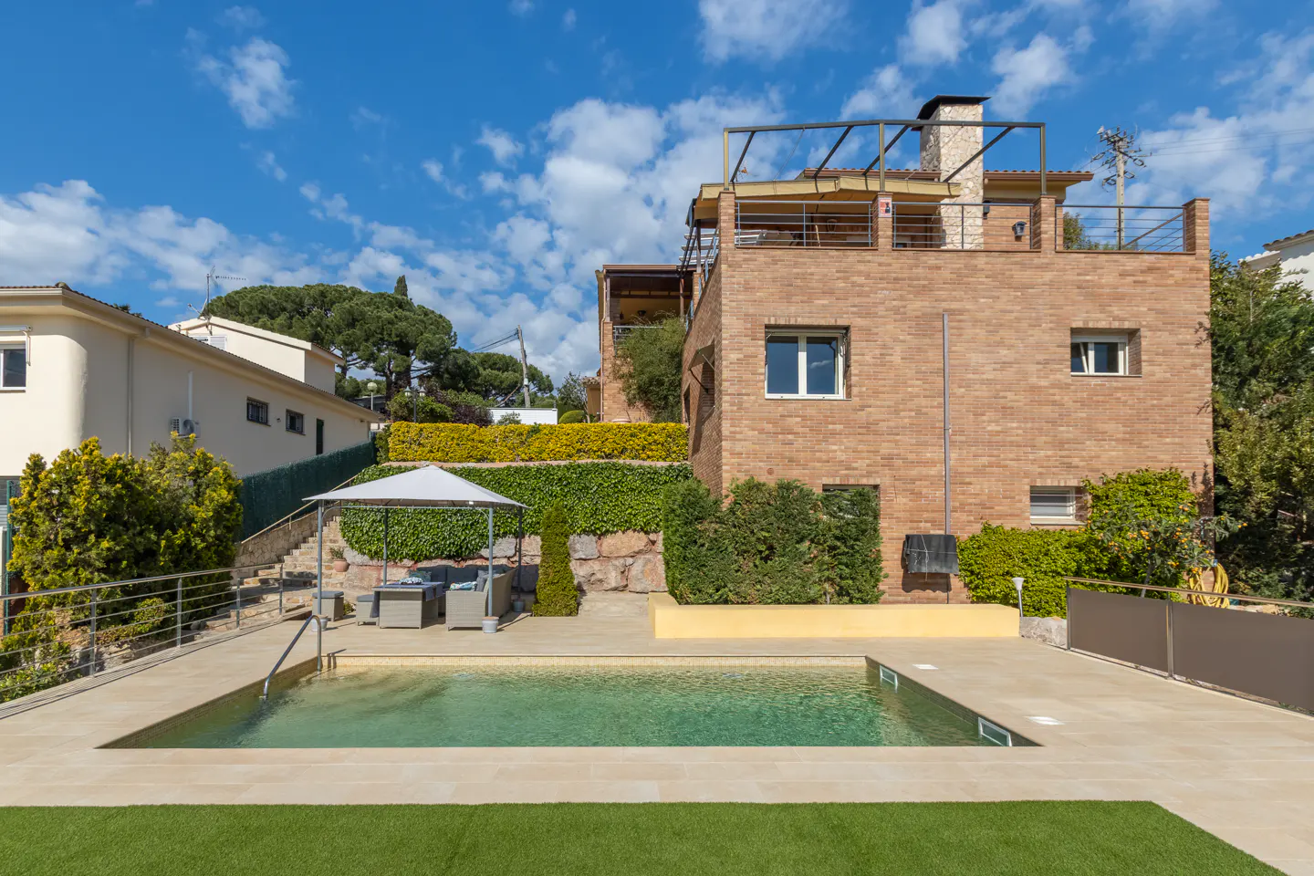Backyard view of a brick house with a pool, patio furniture under a white canopy, and green landscaping on a sunny day.