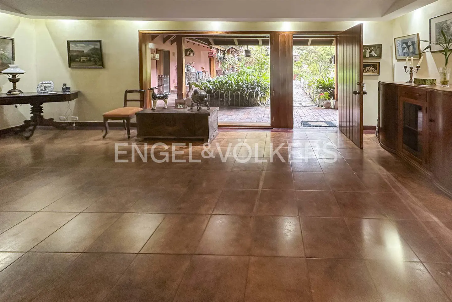 A living room with brown tile floors, a wooden chest, and an open doorway to a garden.
