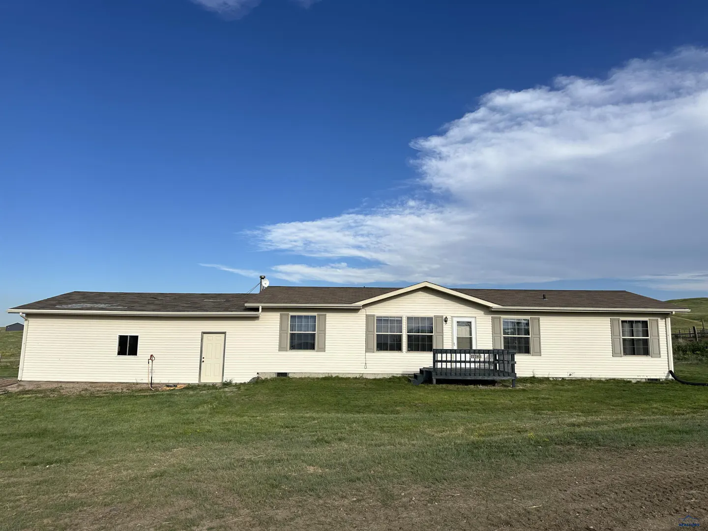 Single-story cream-colored ranch house with a brown roof, small front deck, and green lawn under a bright blue sky.