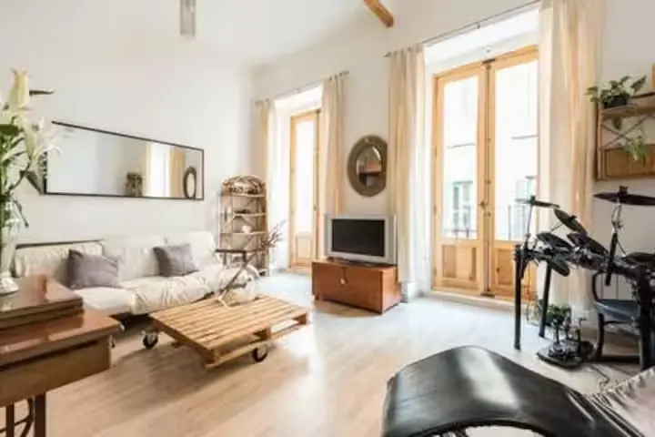 Bright living room with a white sofa, wooden pallet coffee table, television, and a drum set near large glass doors.
