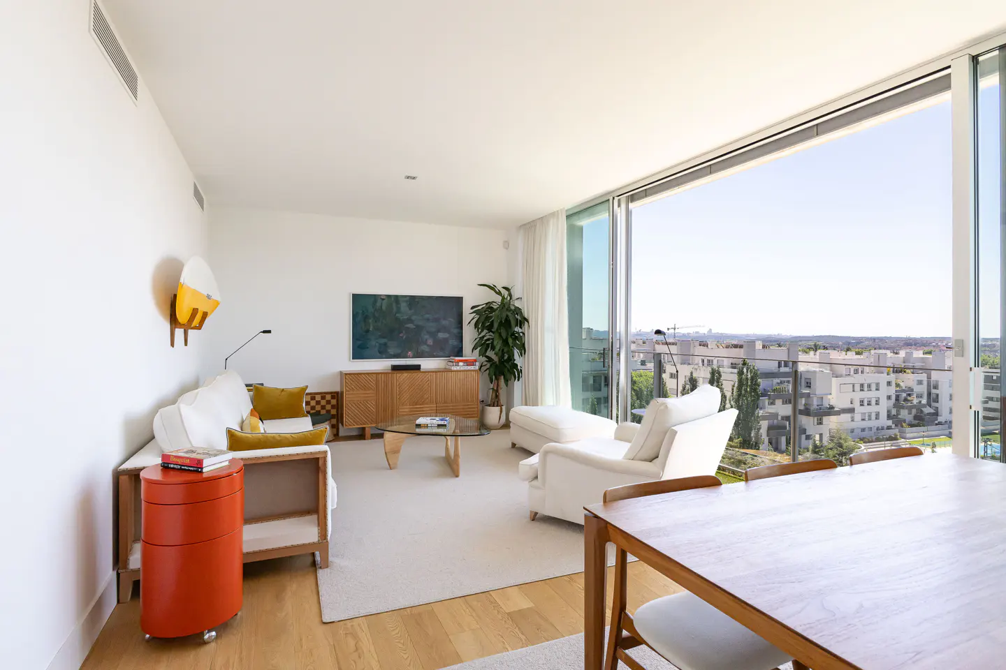 Bright, modern living room with white sofa, wood accents, and floor-to-ceiling windows overlooking a cityscape.