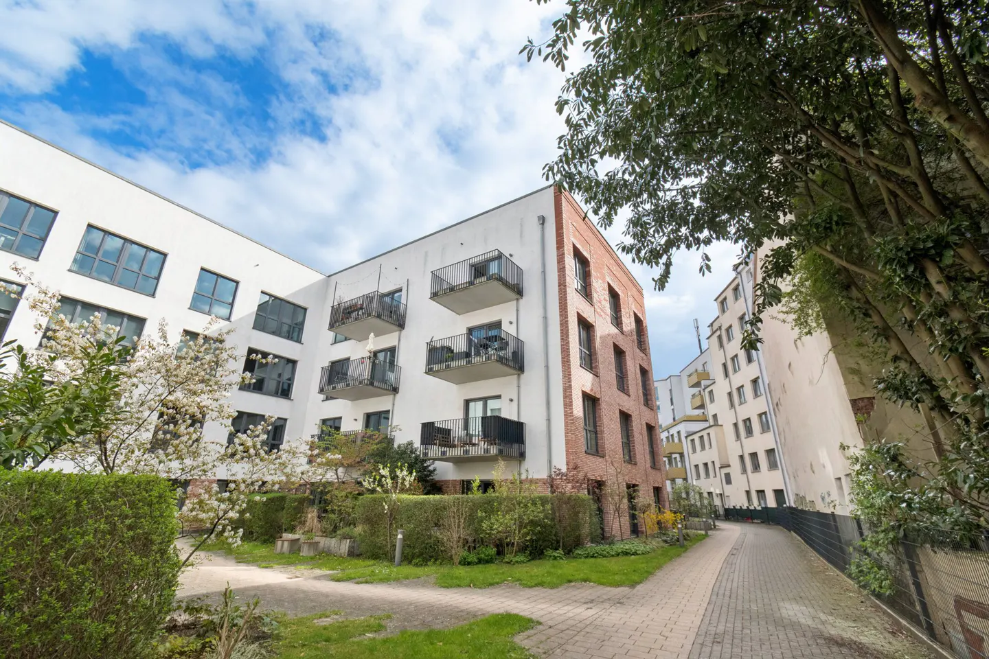 Modern apartment building with white and brick facade, black balconies, and landscaped courtyard with trees and a brick walkway.