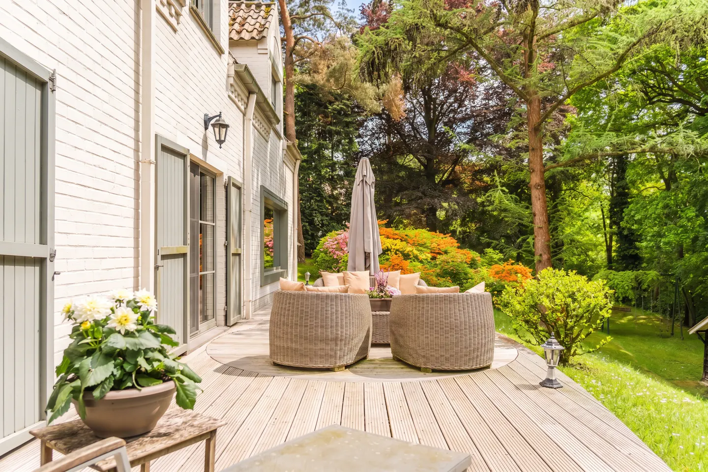 Wooden patio with wicker furniture and umbrella, next to a white brick house with green shutters, overlooking a lush garden.