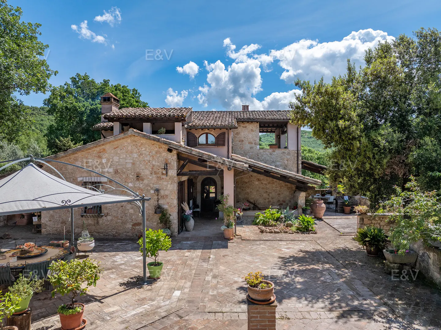 Rustic stone villa with a tiled roof, featuring a paved courtyard, potted plants, and a gazebo under a sunny blue sky.