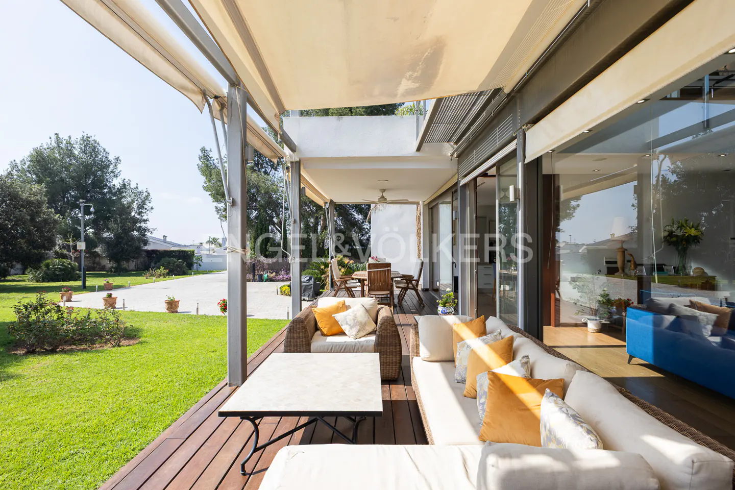 Covered patio with wicker furniture, white cushions, and yellow pillows. A table and chairs are in the background. Green lawn and trees are visible.