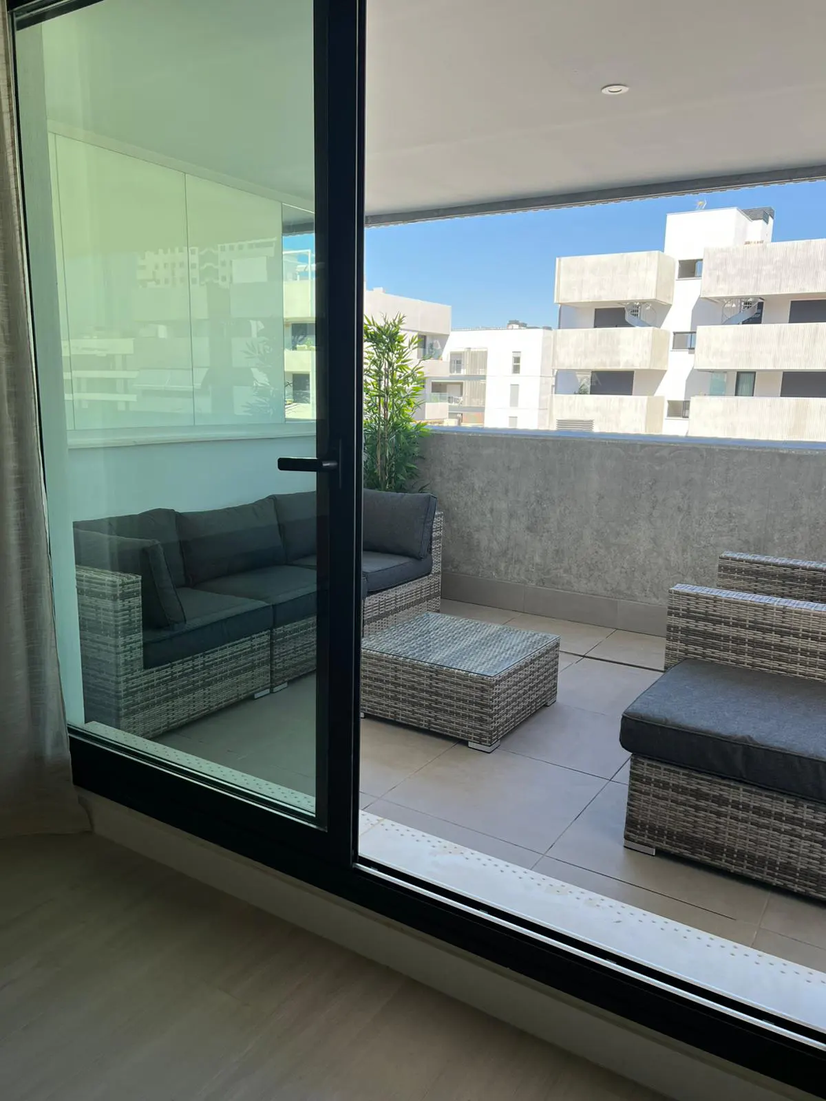 View from inside to a balcony with gray wicker furniture, including a sofa, table, and lounge chair, with buildings in the background.