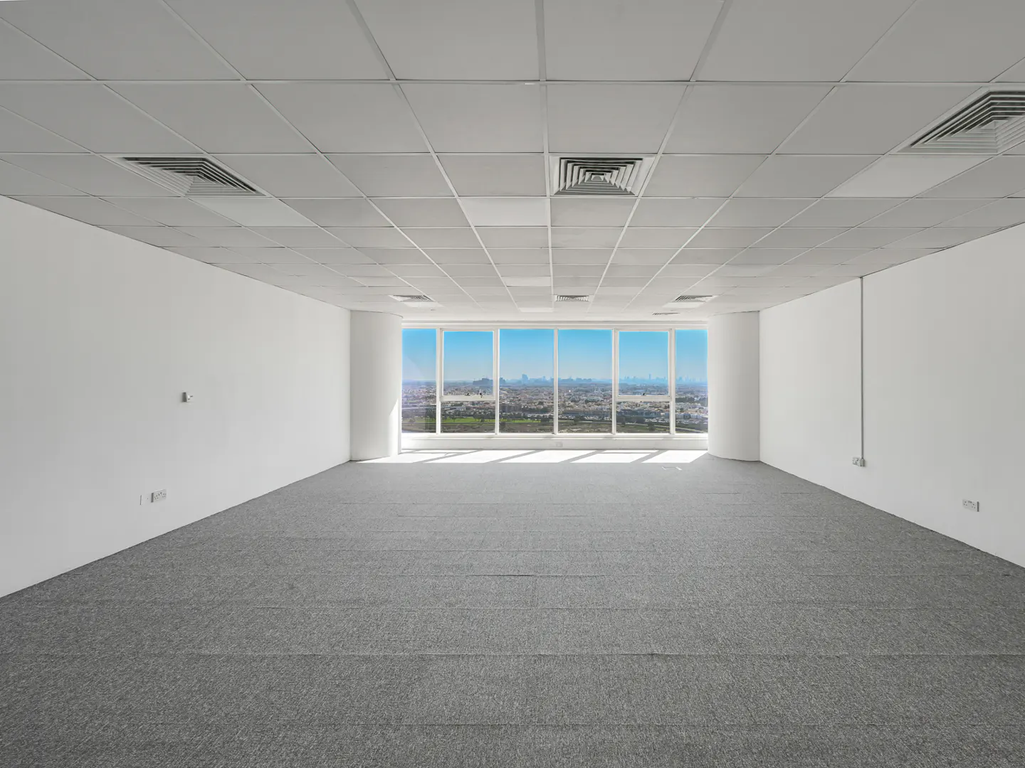 Empty office space with gray carpet, white walls, and a large window showing a city skyline under a blue sky.