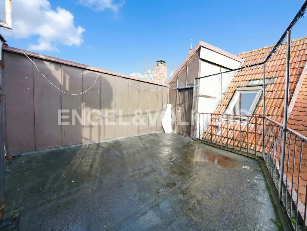 A rooftop terrace with dark flooring, a metal wall, and a sloped roof with terracotta tiles under a clear blue sky.