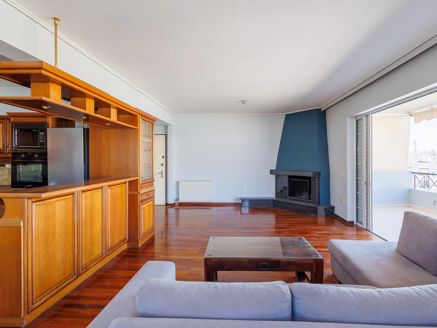 Open-plan living room with wood floors, a light grey sofa, a coffee table, and a kitchen area with wooden cabinets.