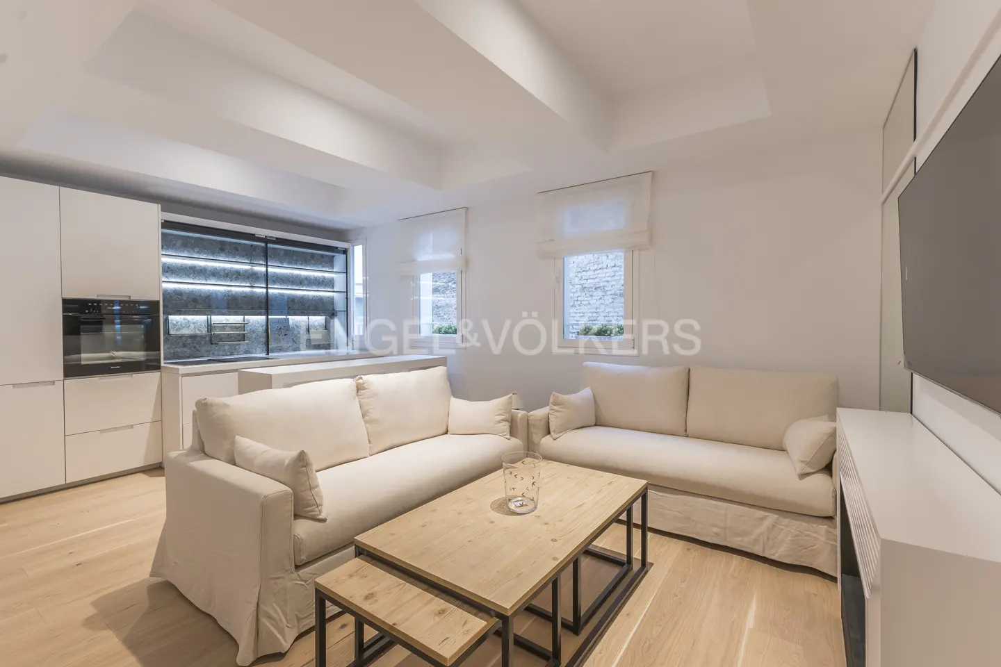Bright, modern living room with two cream sofas, a wooden coffee table, and light wood floors. White kitchen cabinets are visible in the background.