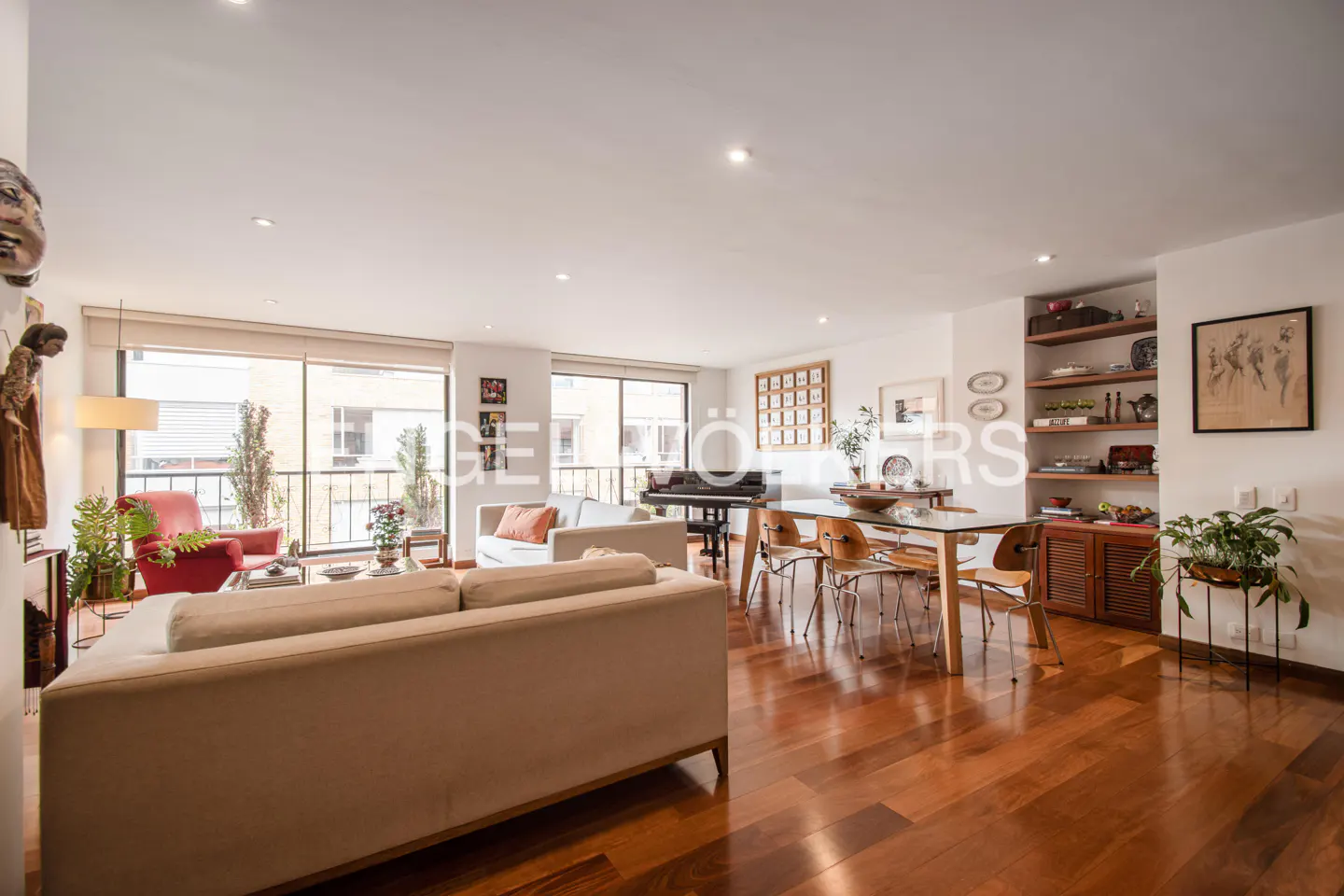 Bright living room with hardwood floors, beige sofas, a dining table, and a piano near large windows. Shelves and artwork decorate the walls.