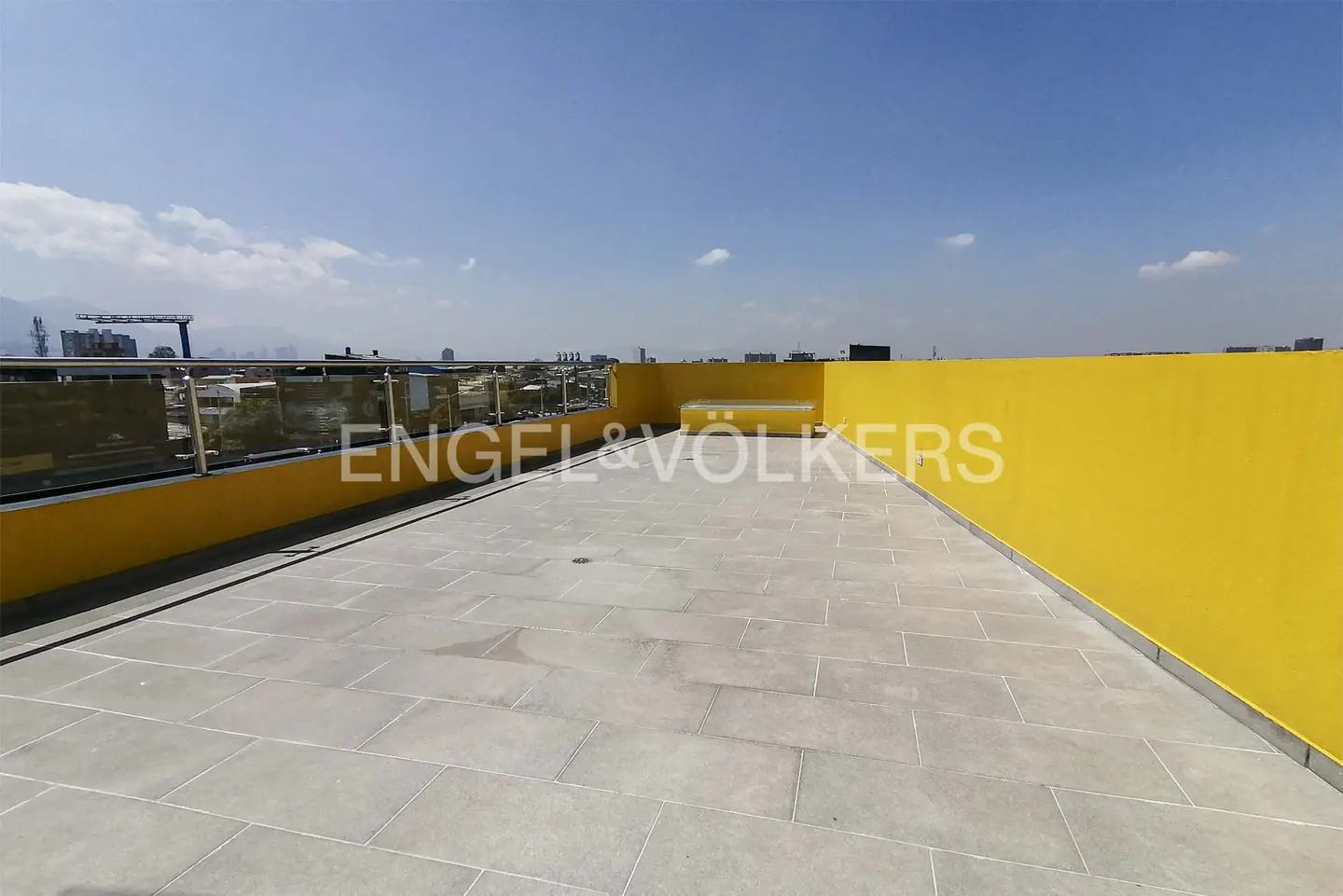 Rooftop patio with gray stone tiles, yellow walls, and glass railings overlooking a cityscape under a blue sky.