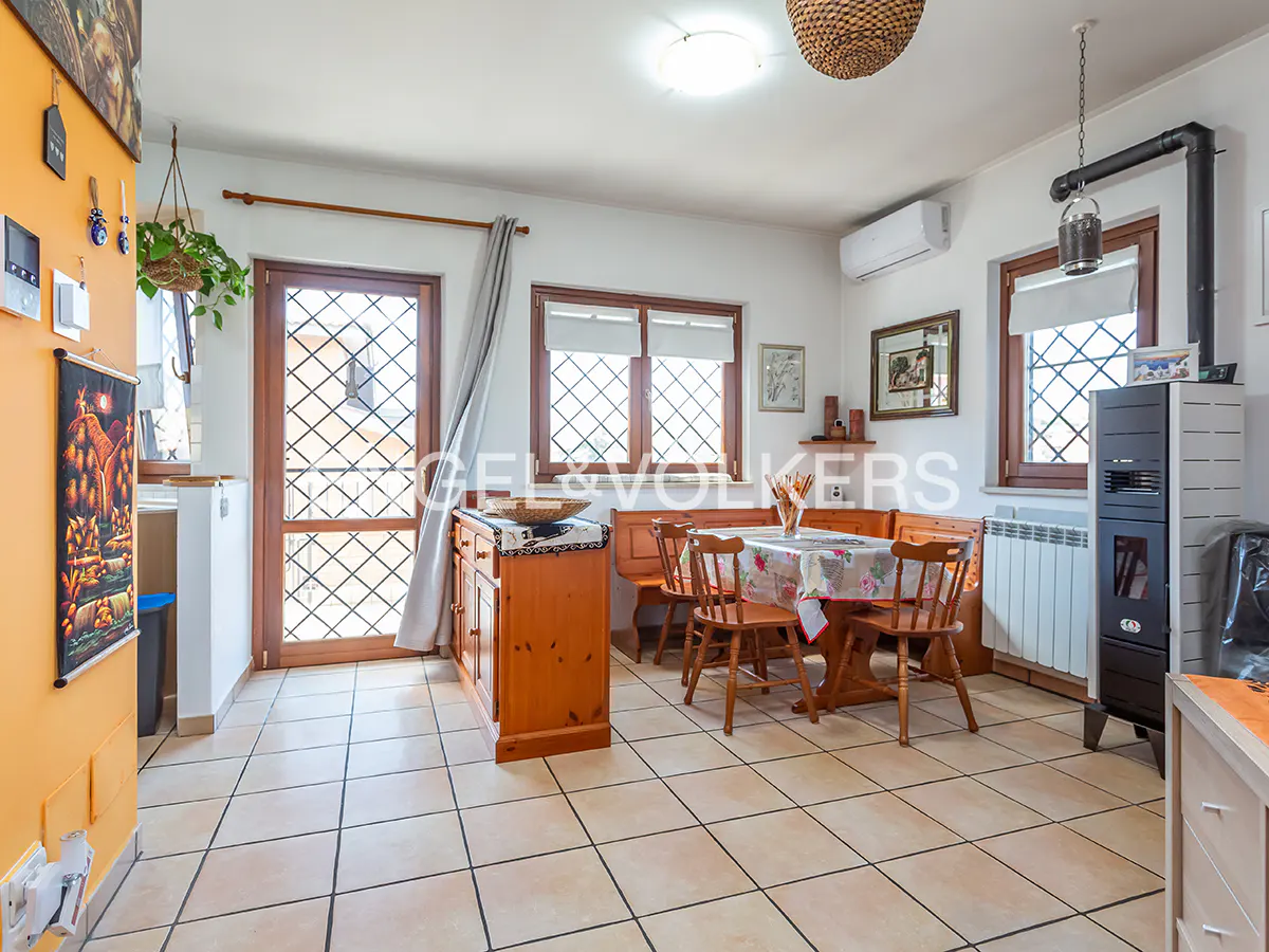 A bright dining room with a wooden table, chairs, and a bench. A wood-burning stove stands near a window. Tiled floor and orange wall.