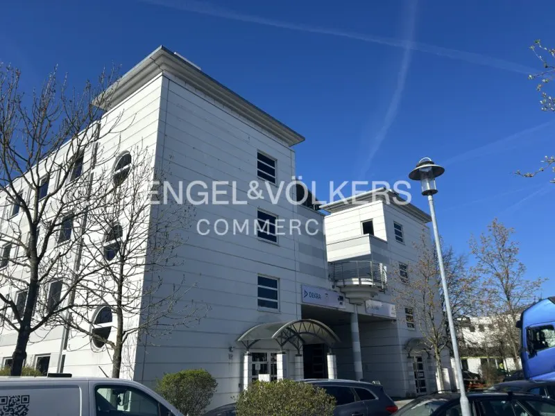 Exterior of a white, multi-story commercial building with Engel & Völkers signage under a blue sky. Cars and trees are in the foreground.