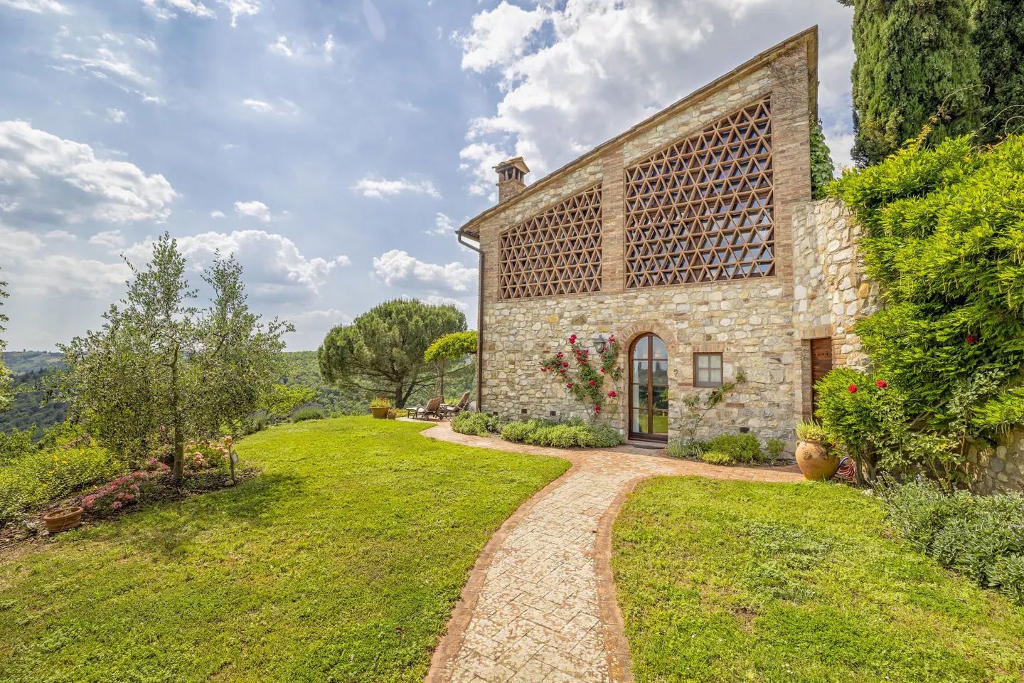 A stone villa with a triangular lattice facade, arched doorway, and brick path set in a lush garden under a blue sky.