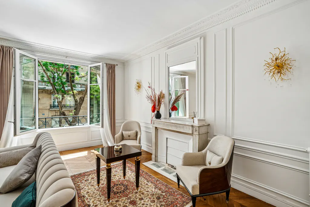 Bright living room with white walls, herringbone floors, and a large open window. Two chairs face a fireplace with a mirror above. A patterned rug and dark wood table sit in the center.
