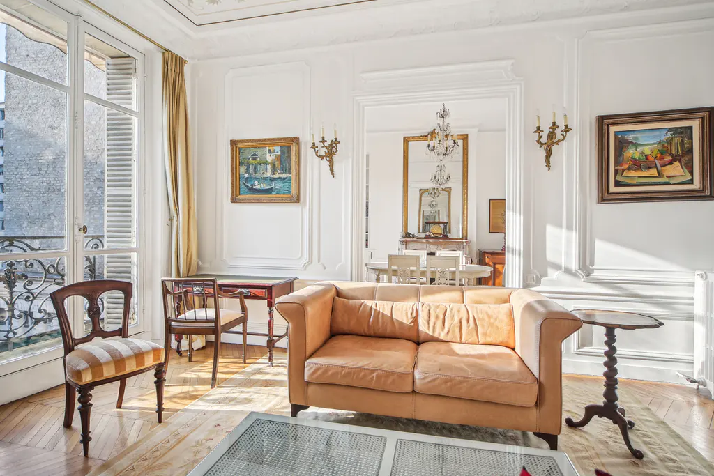 Elegant Parisian living room with a tan leather sofa, antique wooden furniture, paintings, and a balcony view.